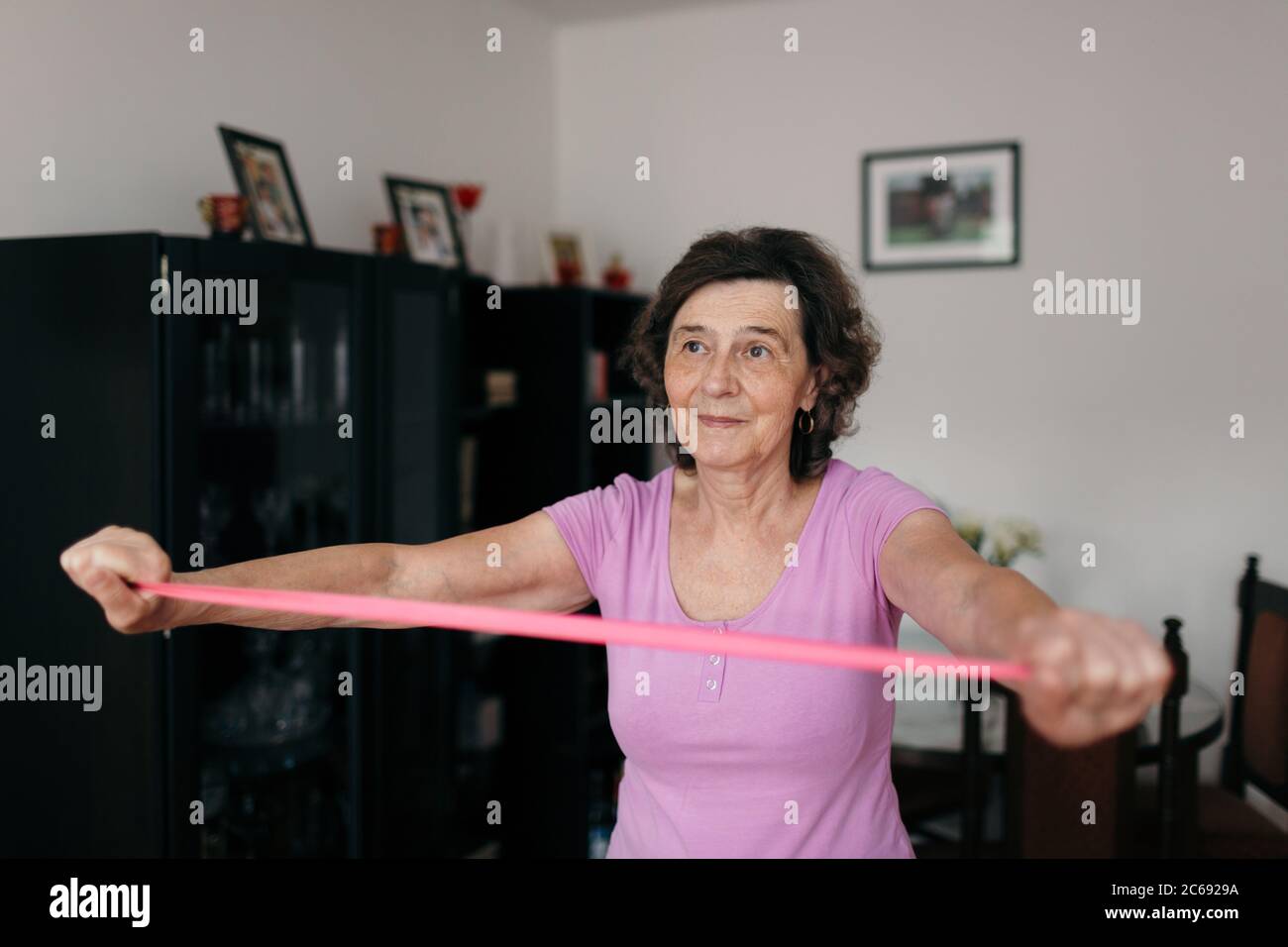 Elderly woman stretching rubber band at home. Waist up shot of 70 years old woman in purple t-shirt exercising arms with pink rubber band in living ro Stock Photo