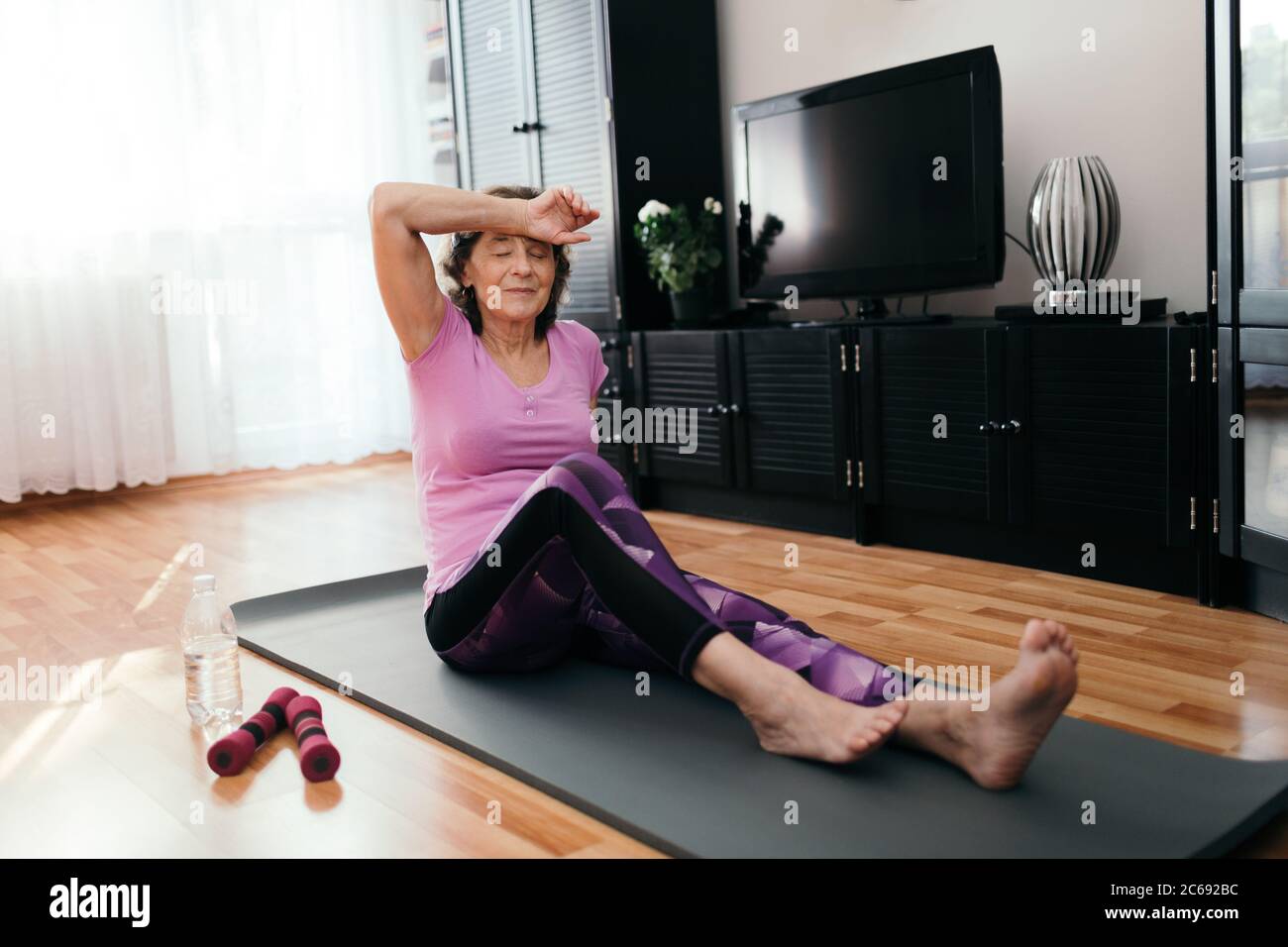 Tired elderly woman after exercise at home. Weary senior woman in purple sportswear sitting on fitness mat wipes sweat from her forehead after workout Stock Photo