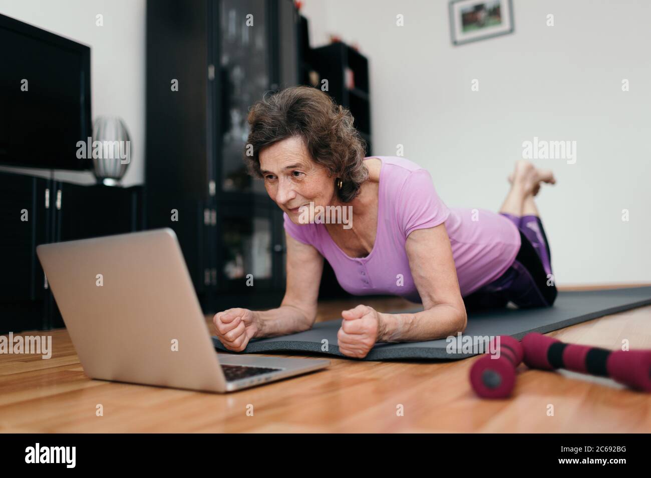Elderly woman exercising at home watching online course. Low angle shot of senior woman at her 70s lying on floor exercising plank. Stock Photo