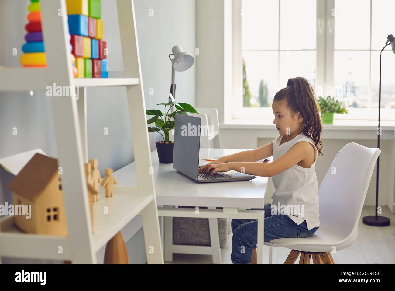 Online education of children. Serious little girl is typing laptop text while sitting at a table at home. Stock Photo