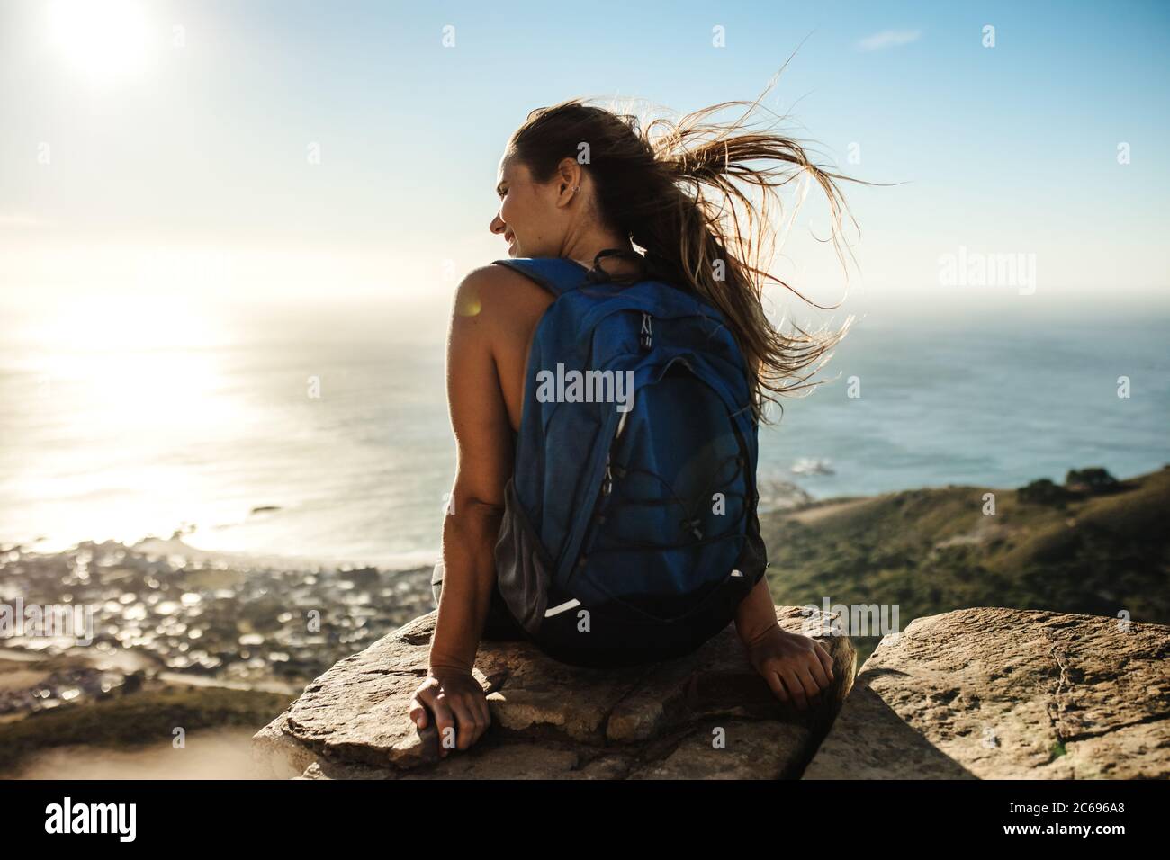 Rear view of a woman with backpack sitting on the cliff and looking at the view Female hiker relaxing on mountain top. Stock Photo