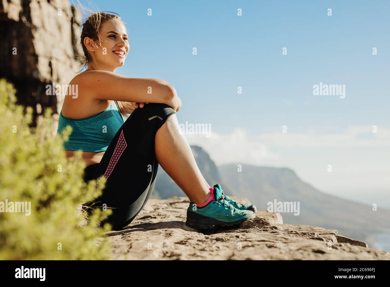 Fitness woman sitting on the cliff looking at the view and smiling. Female trekker resting on mountain top. Stock Photo