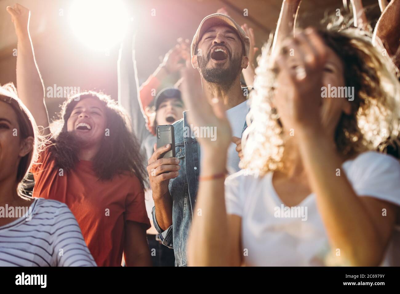 Large group of football fans in stadium yelling and cheering with excitement for their team. Excited sports fans standing with their hands raised and Stock Photo