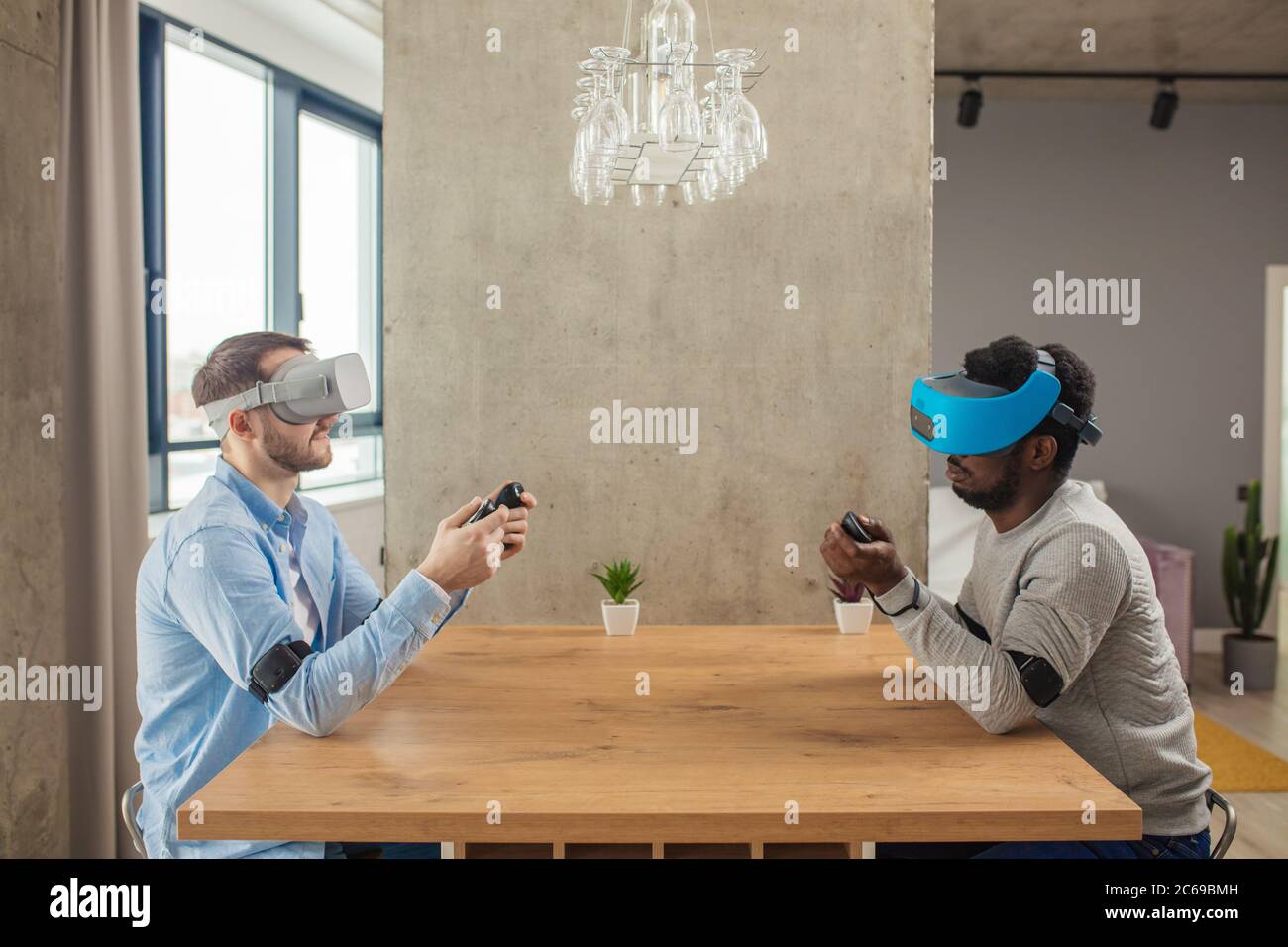 Cheerful interracial male gamers having fun with new trends technology, sitting against each other at table with virtual reality headset on . Stock Photo