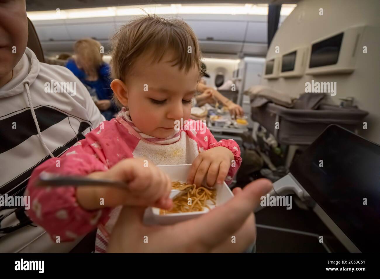 little cute hungry baby sitting in an airplane on his parents lap and eating delicious special food on board with a fork Stock Photo