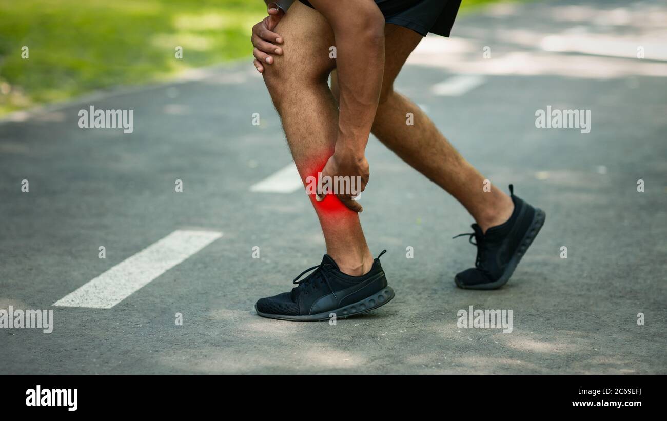 Millennial runner holding his injured ankle on road outdoors, closeup