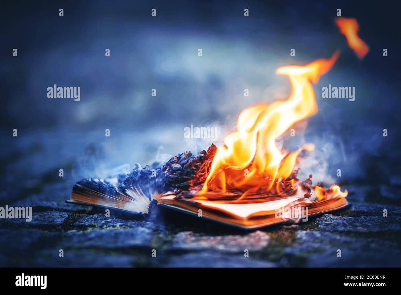 An old book lies on a rocky path made of cobblestones and its pages burn with a bright strong flame and smoke blue smoke at night. Magic. Censorship. Stock Photo