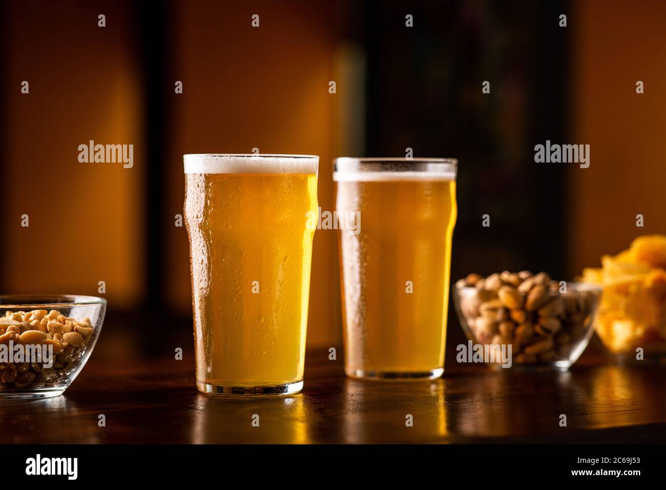 Two glasses of lager on wooden table, near are pistachios, nuts and chips in glass plates Stock Photo