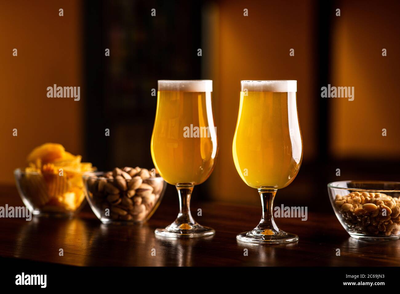 Two glasses with light beer and foam on wooden bar counter in interior of pub, pistachios, crackers and nuts in glass plates nearby Stock Photo