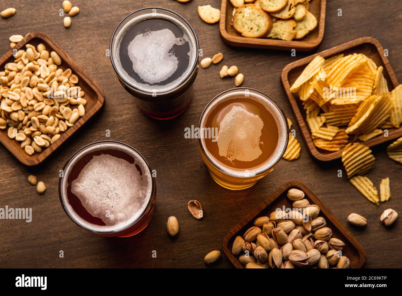 Craft drinks in glasses and pistachios, nuts, crackers, crisps and chips in plate and scattered on wooden table Stock Photo