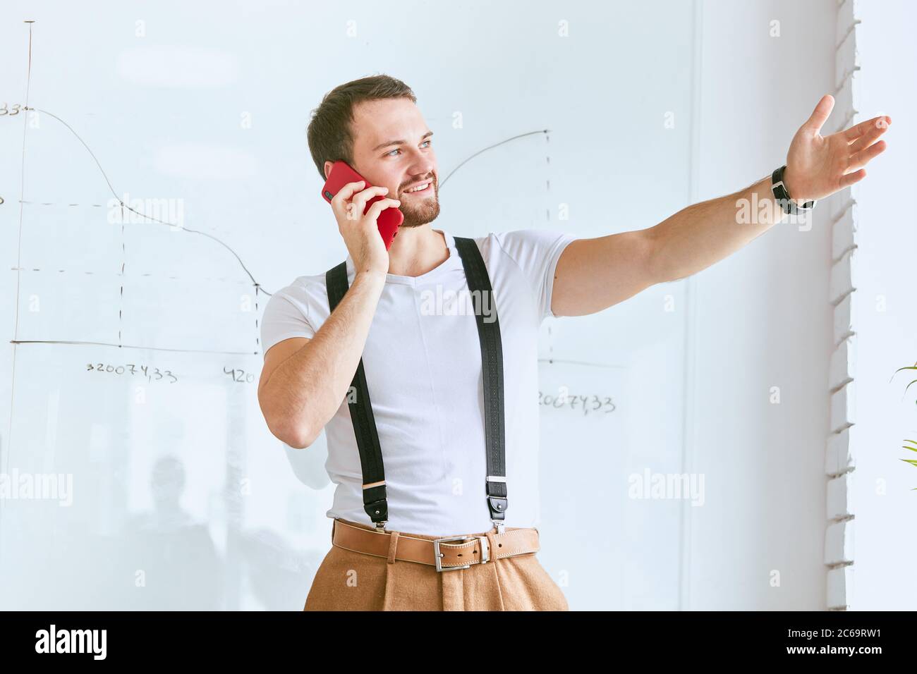 Young businessman wearing white t-shirt and trousers with suspenders look side, talking on phone with business partners. Chart diagrams background Stock Photo