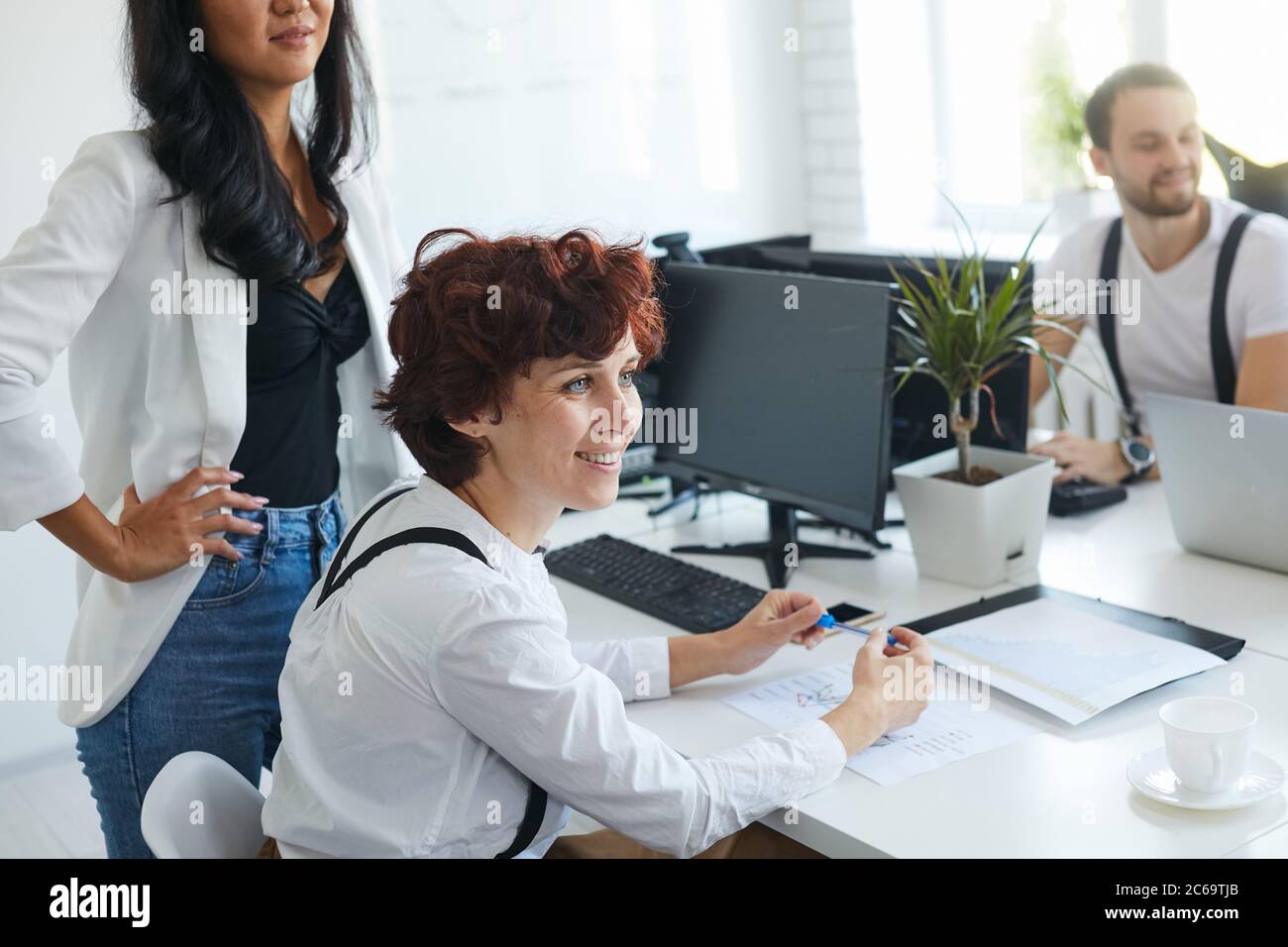 Beautiful smiling woman look side at coach, attentively listen to speaking,colleagues around her, sit on table with computers, papers Stock Photo