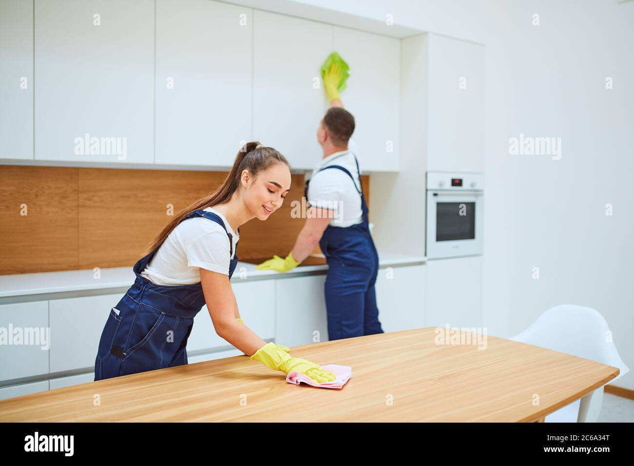 Young professionals of cleaning service do spring-cleaning in kitchen. Woman in special equipment wiping dust off from table, background man wiping du Stock Photo
