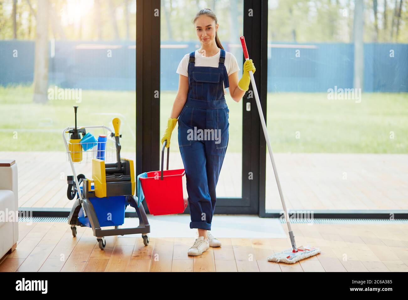 Caucasian woman holding red bucket for floor washing and other equipment for cleaning house. Staff of cleaning service, panoramic window background Stock Photo