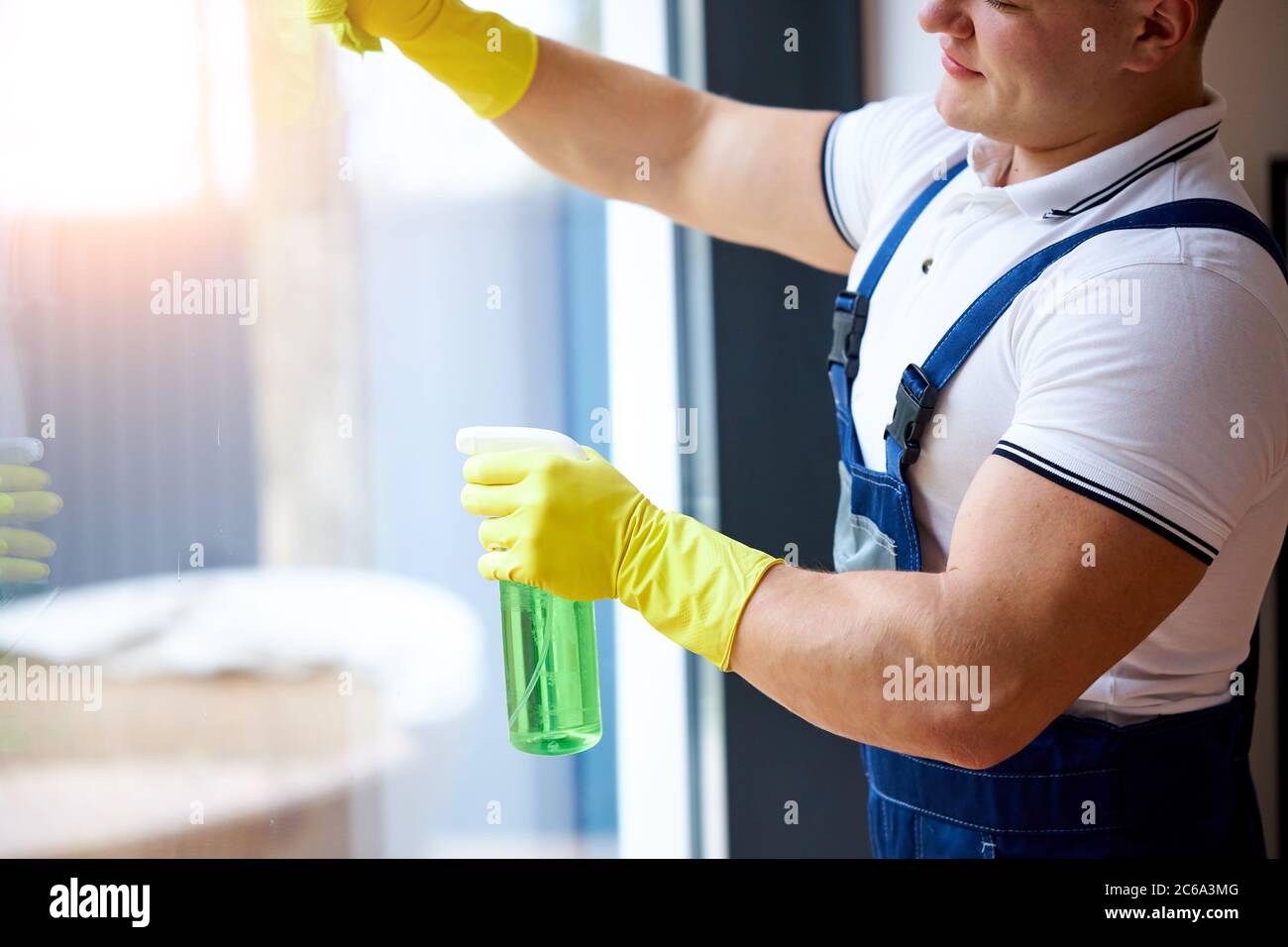 Young muscled guy in uniform from cleaning service wearing yellow protective gloves, holding spray wash the window Stock Photo
