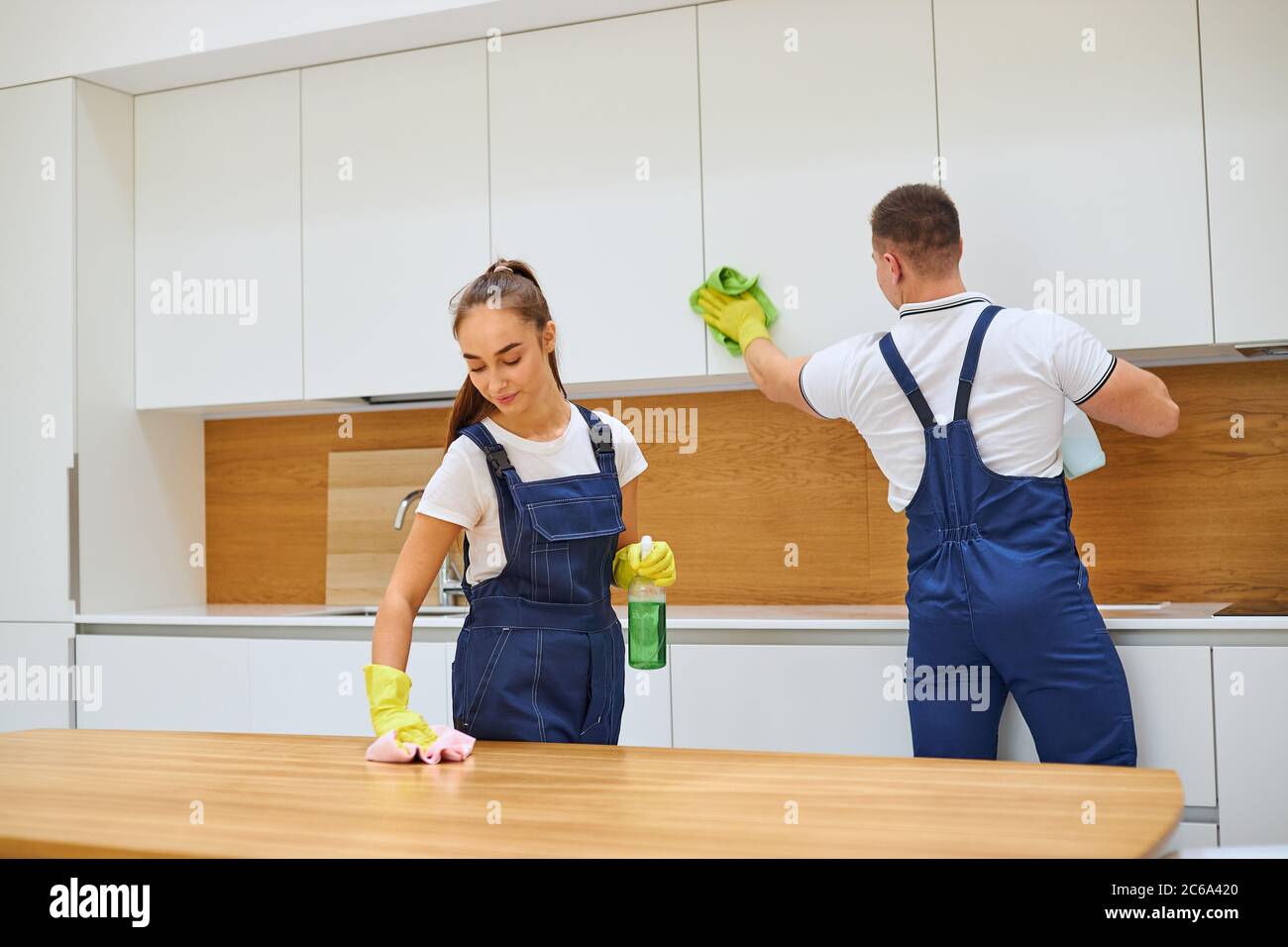 Young caucasian cleaners in uniform cleaning kitchen together, wiping dust off wooden table. White interior Stock Photo