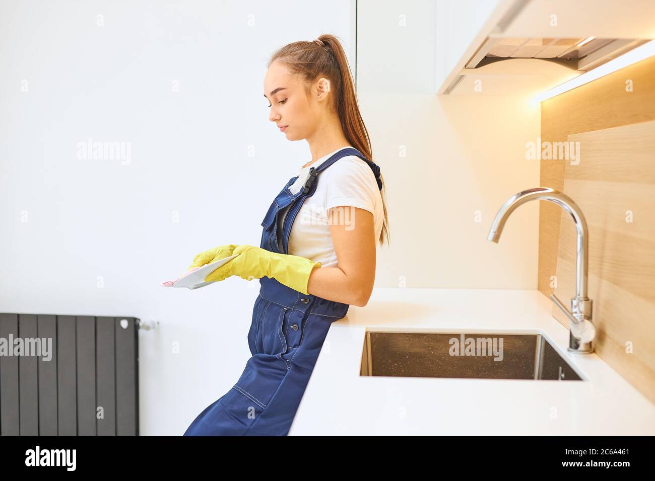 Side view on beautiful caucasian janitor in blue uniform stand wiping dishes in kitchen, white interior of kitchen. Young cleaner wearing yellow rubbe Stock Photo