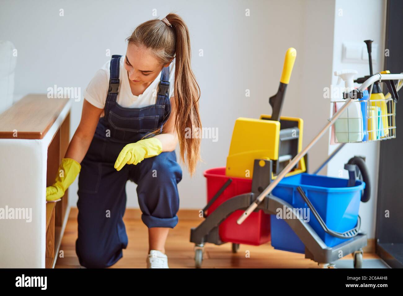 Young staff of cleaning room. Beautiful girl wearing special equipment with yellow rubber gloves, using spray wiping dust off. Careful worker of clean Stock Photo