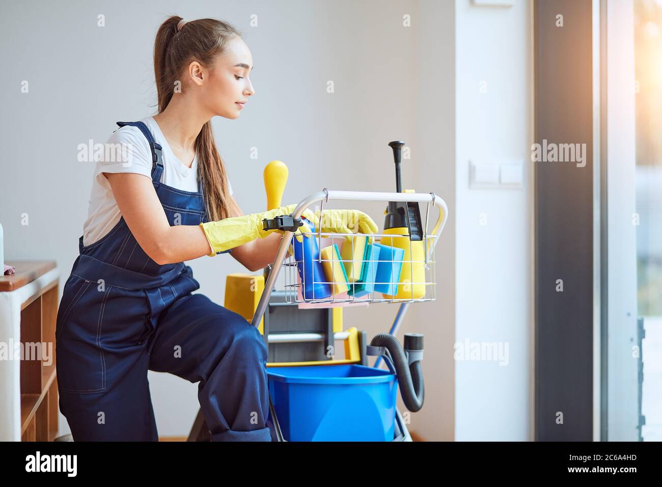 Young beautiful staff of cleaning company in special clothes, equipment. Wearing rubber gloves, sit opposite window Stock Photo