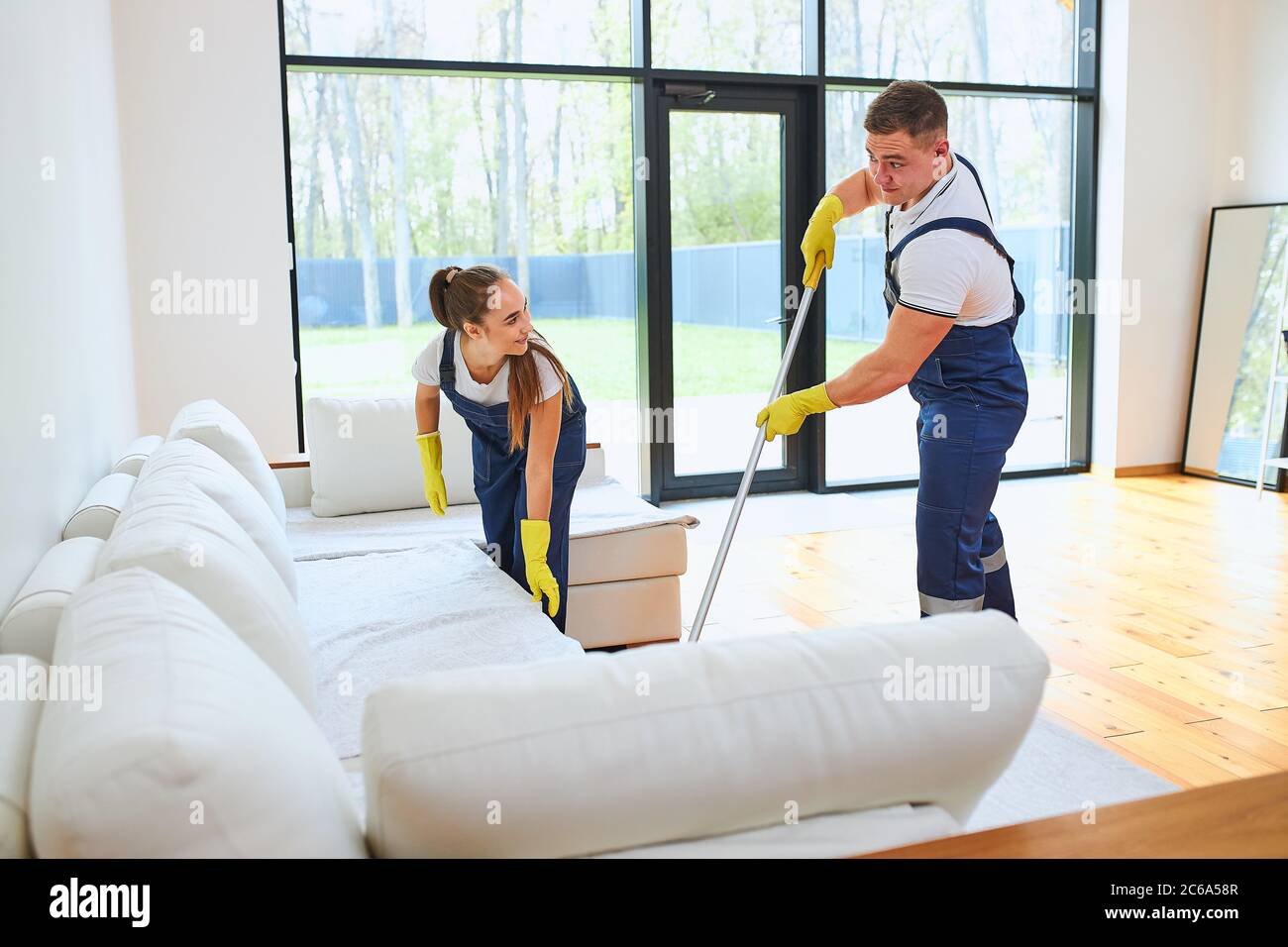 Two janitors in uniform cleaning new room with white sofa, mopping floor, straighten sofa Stock Photo