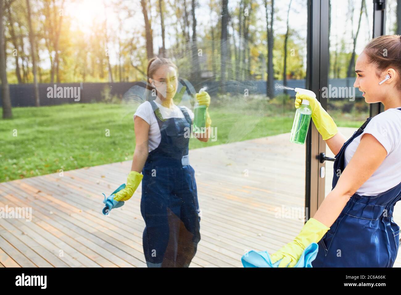 CLeaning service girl in uniform wearing yellow protective gloves washing window outdoors, look at reflection of herself in glass Stock Photo