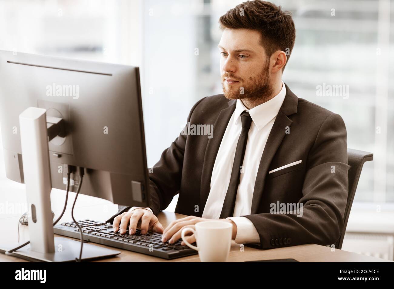 Business concept. Successful young businessman at work. Manager sitting at the office table and working on computer. Busy man in suit indoors on glass Stock Photo