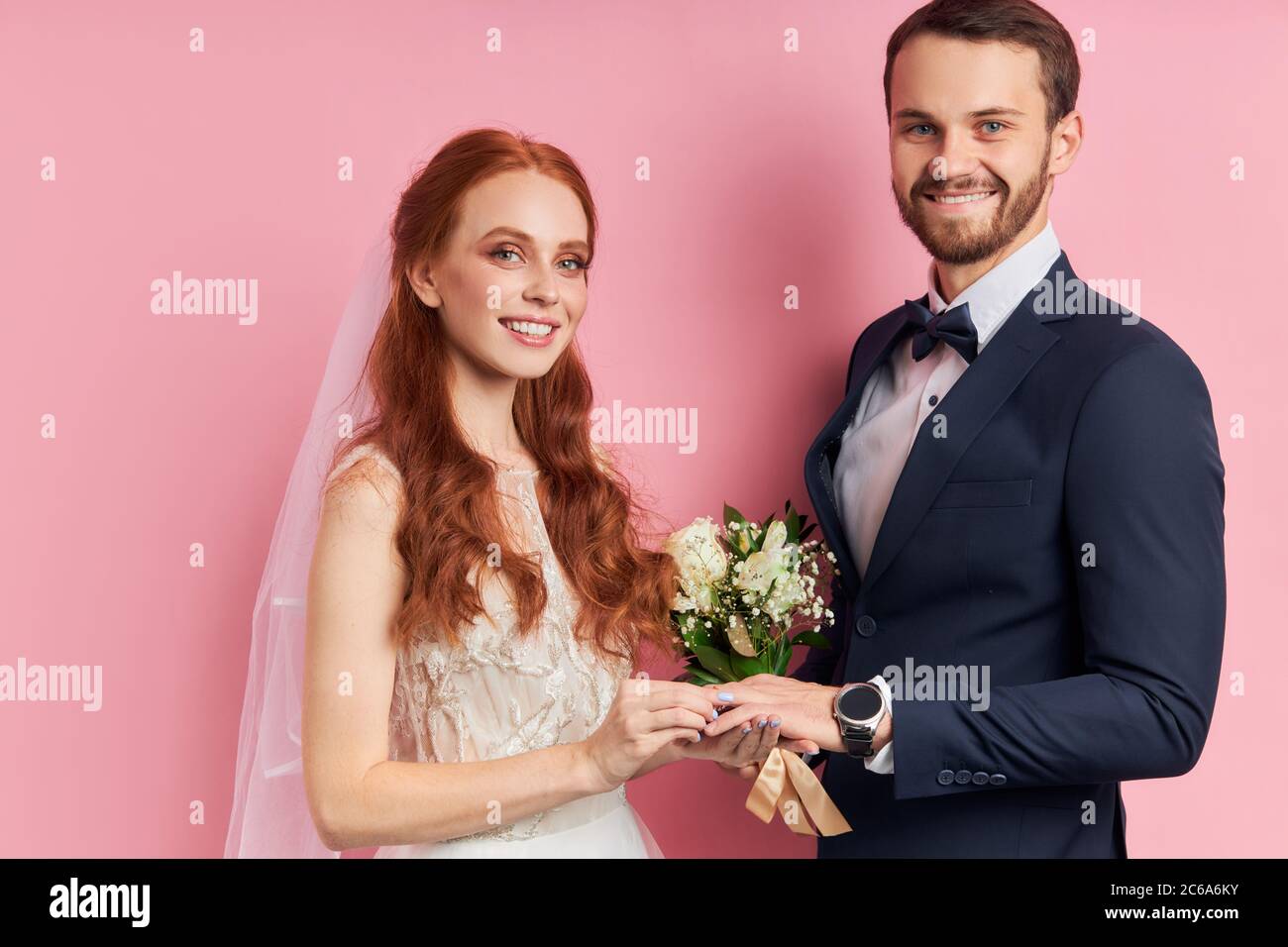 Happy male in suit and female in white wedding dress putting golden ring with diamond on finger, stand isolated over pink background. Love, marriage c Stock Photo