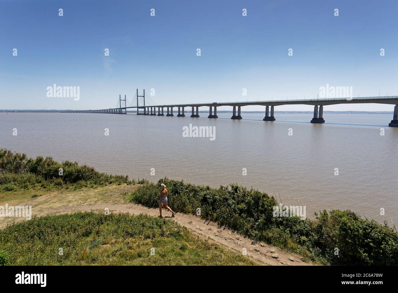 A woman walks along the path which runs alongside the river Severn estuary which overlooks the Prince of Wales Bridge as seen from Sudbrook in Monmout Stock Photo