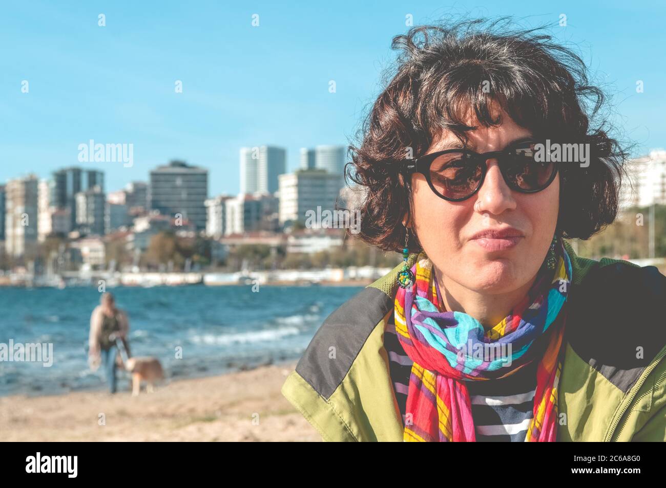 Outdoor portrait of a pretty curly woman with colorful clothing and sunglasses, sitting on the beach with a sea background. Urban lifestyle. Stock Photo
