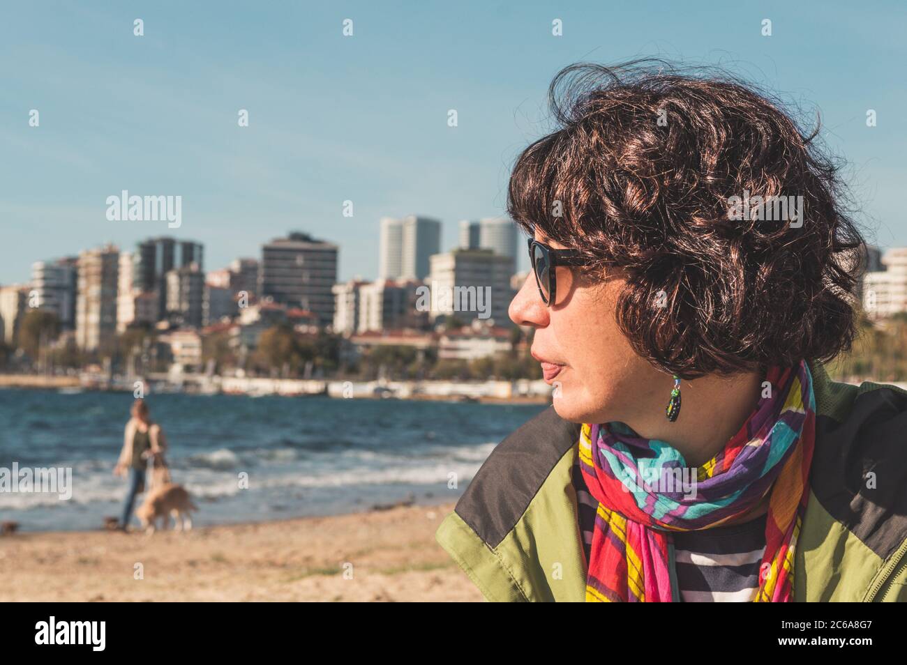 Profile of a brunette woman with curly hair, colorful clothing, and sunglasses, sitting on a sandy beach and looking away. Urban lifestyle. Stock Photo