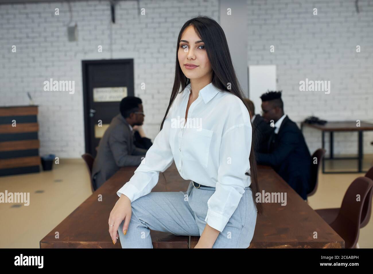 Cute young woman wearing white formal shirt sit on table in office, confidently looking at camera Stock Photo
