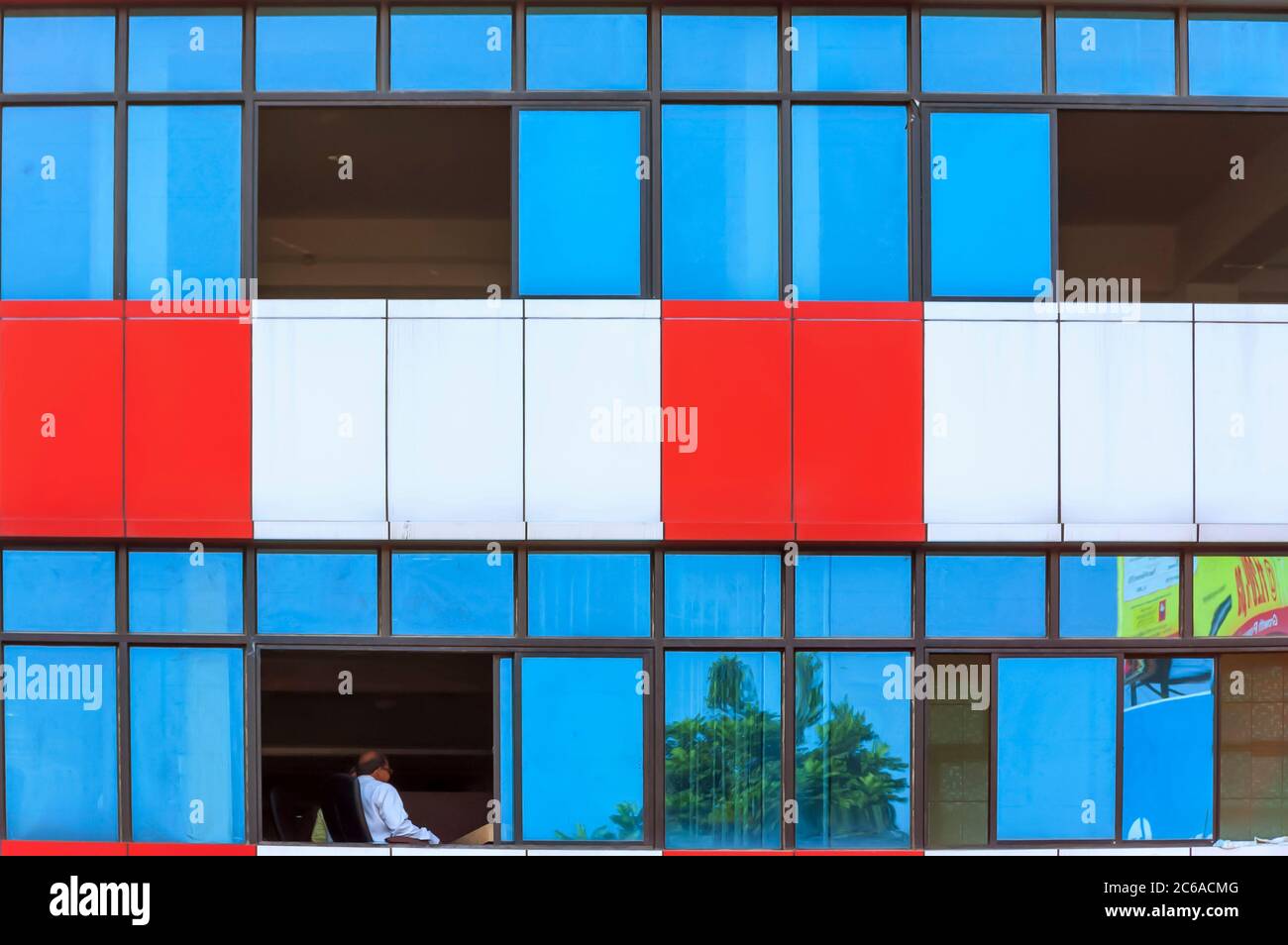 A figure of a man sitting on a chair visible through an open window of a multicolored modern building in Guwahati, Assam, India. Stock Photo