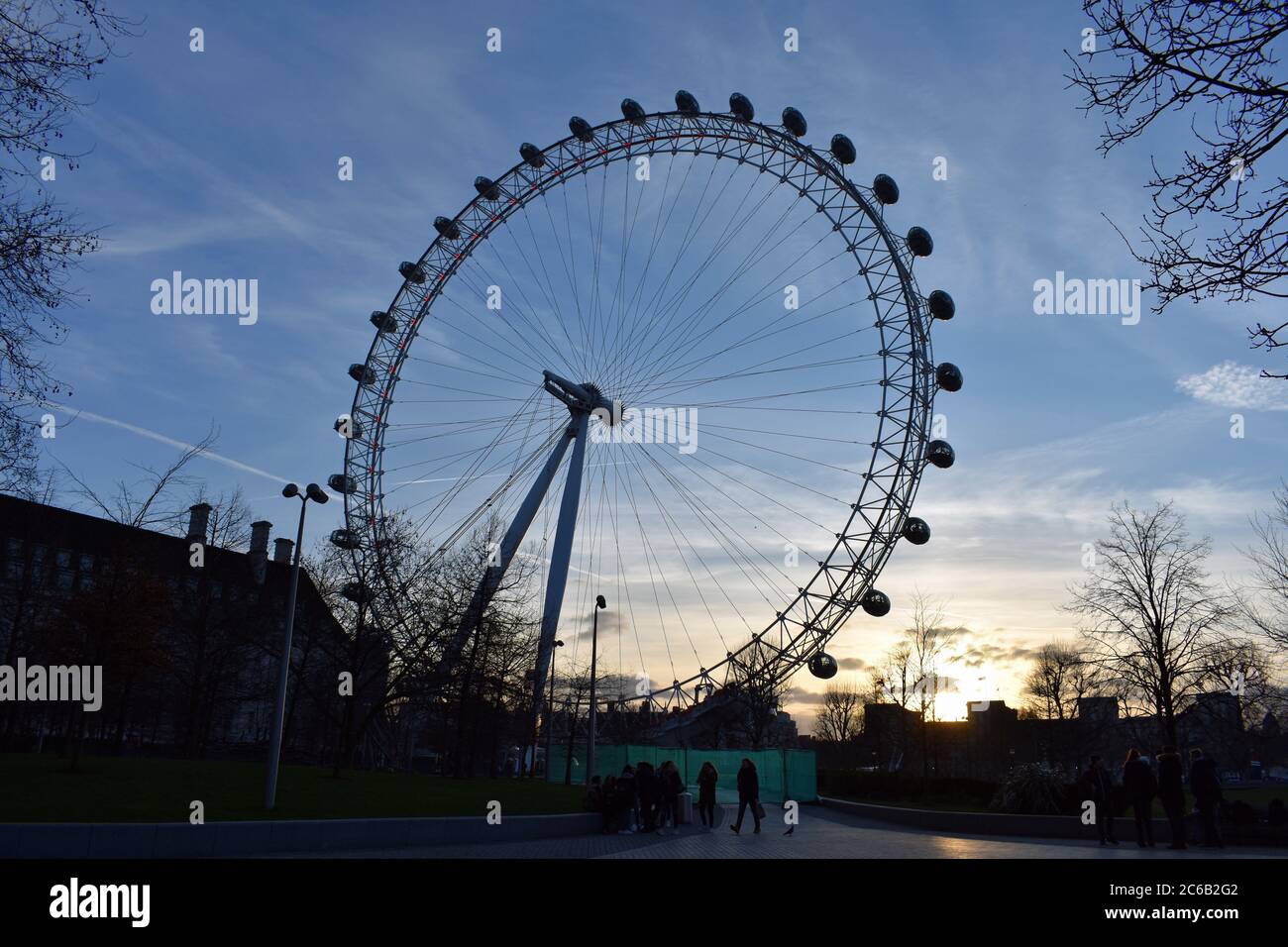 The London Eye at sunset along the South Bank of the River Thames.  A group of people can be seen by a green temporary fence below the wheel. Stock Photo