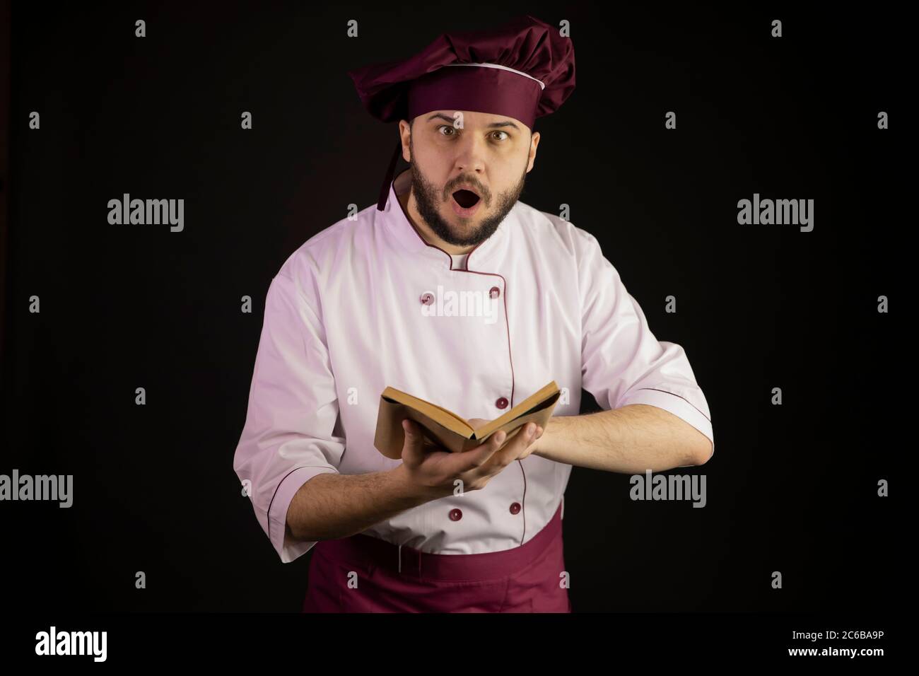 astonished bearded Master chef in uniform keeps open book of vintage old recipes Stock Photo