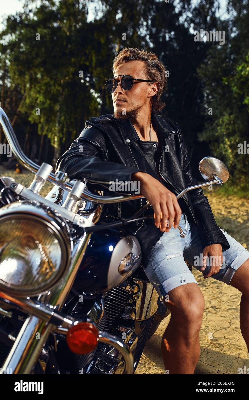 Brutal young man in sunglasses, blue jeans and a black leather jacket  sitting on the custom motorcycle Stock Photo - Alamy