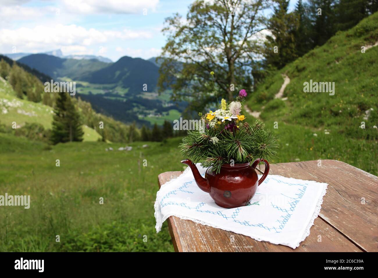 Old tea pot with wild flowers bouquet on wooden table. Blurred background of lush meadows and Alps mountains. Hintersee valley, Berchtesgadener Land Stock Photo
