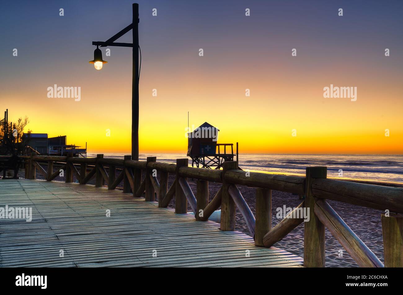 Sunrise view from a walking street and the sun behind the silhouette of the streetlight and the lifeguard box in the beach. Stock Photo