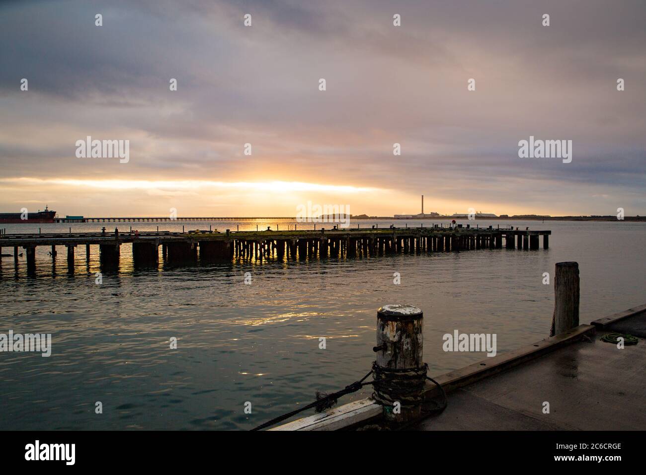 The sun rises through the clouds over the calm sea and wharves in Bluff, New Zealand Stock Photo