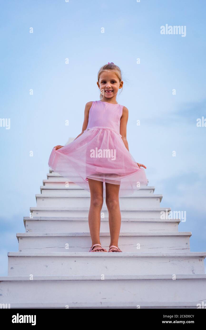 A girl in a pink dress stand on a white staircase extends into blue sky ...