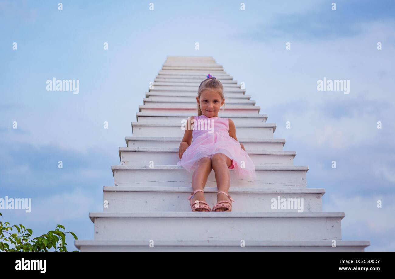 A girl in a pink dress sit on a white staircase extends into blue sky ...