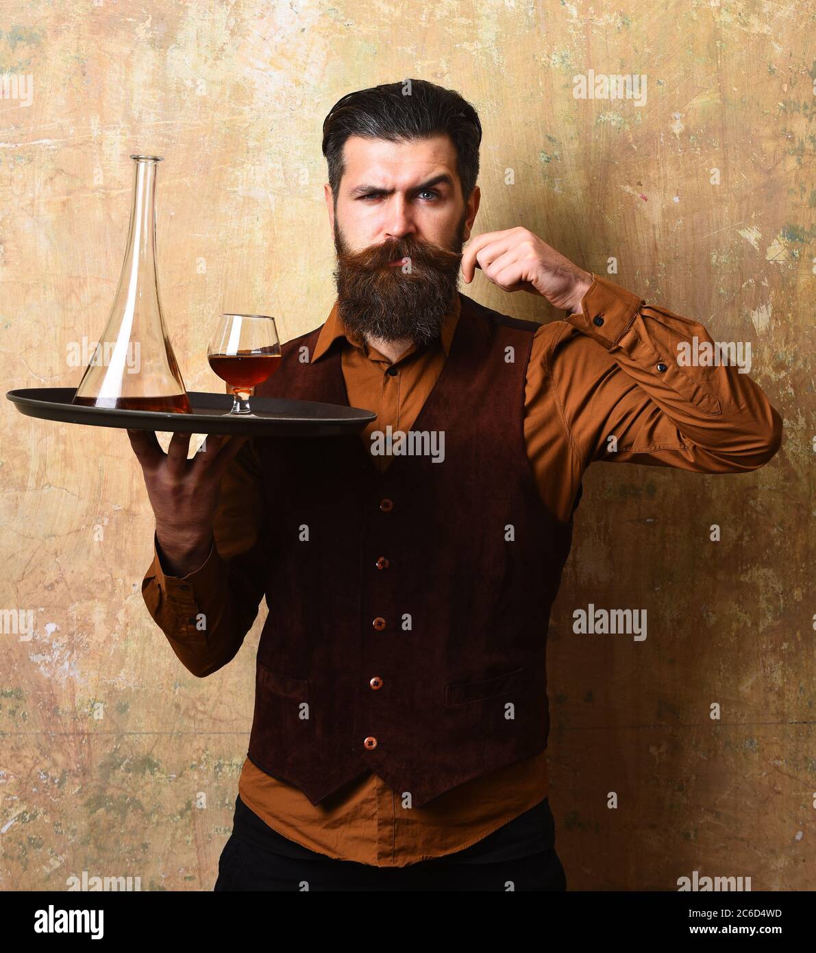 Barman with confident face serves scotch or brandy. Man with beard curls mustache holding cognac on beige wall background. Waiter with glass and bottle of whiskey on tray. Service and drinks concept Stock Photo
