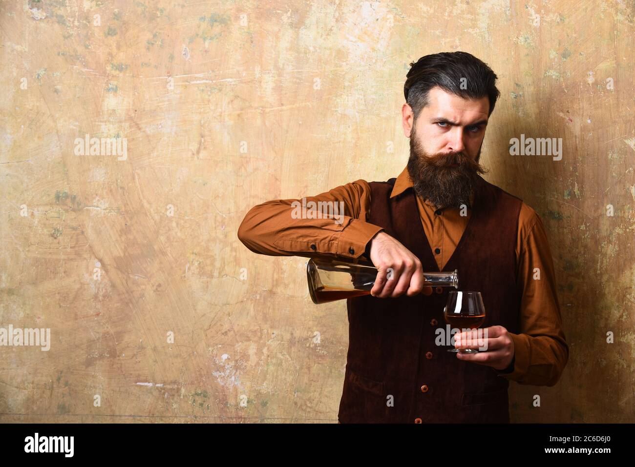 Guy with glass and bottle of cognac. Man with beard and mustache pours alcoholic beverage on beige background. Macho with strict face drinks brandy or whiskey. Service and restaurant drinks concept Stock Photo