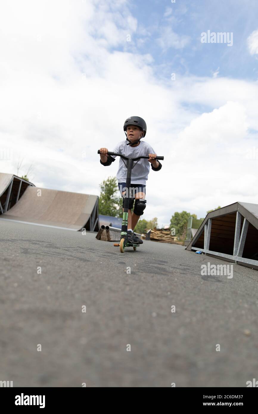 A cute little boy rides a scooter in a skatepark. A young novice athlete spends free time in extreme sports. Lifestyle. Stock Photo