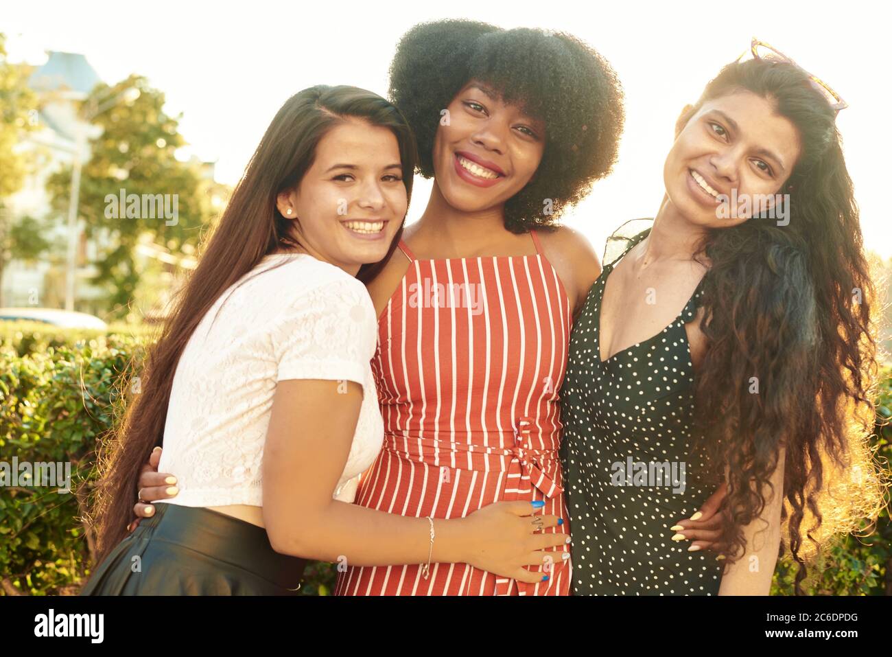 Three close girlfriends or sisters are hugging, standing in the city on a background of trees. The concept of female friendship and youth. Stock Photo