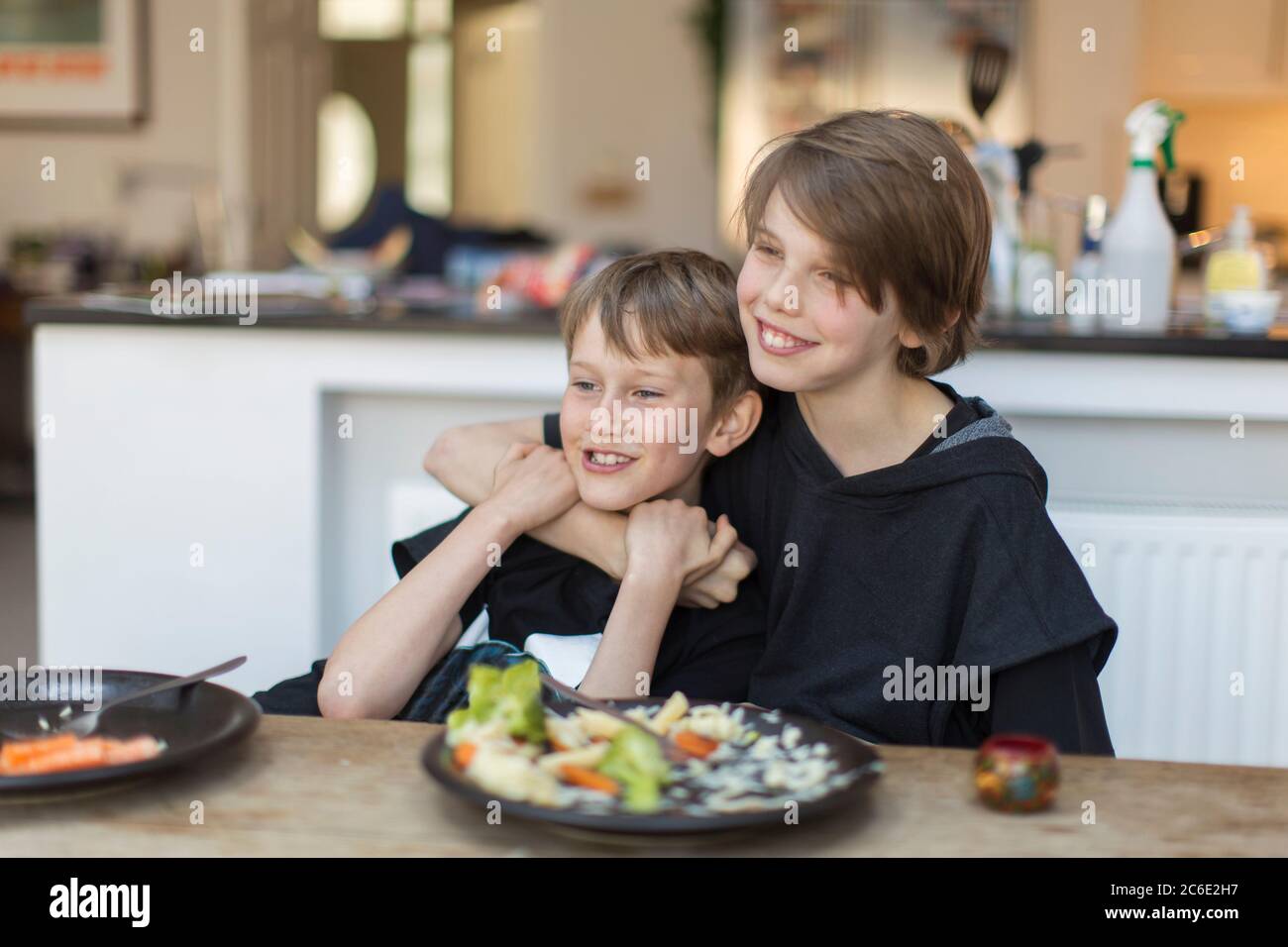 Happy brothers hugging and eating dinner at dining table Stock Photo