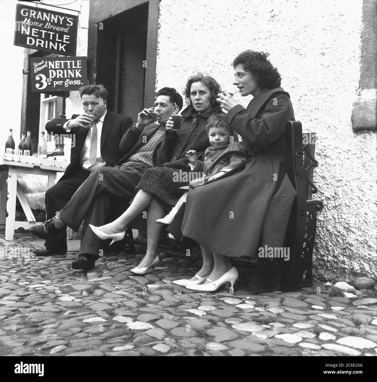 1950s, historical, sitting outside a cottage on a cobbled street at Heysham, Lancashire, England, UK, two men and women drinking 'Granny's Home Brewed Nettle Drink' at 3d per glass. Made from herbal extracts, sugar, yeast, lemons and nettles, the home brew, said to have several health benefits, was made by a local well-known figure in the seaside village, Granny Hutchinson. Stock Photo