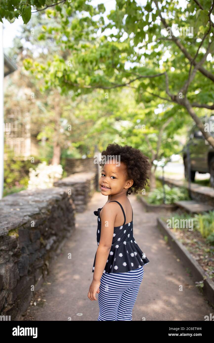 Young beautiful African American girl standing in urban environment while looking back at camera. The background includes a sidewalk and trees. Stock Photo