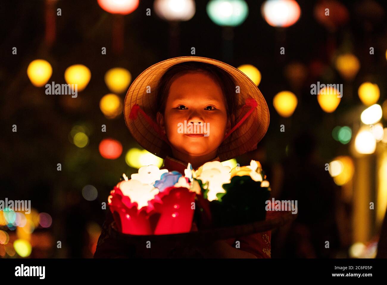 Little Vietnamese girl in hat holding colored paper lanterns with lights behind Stock Photo