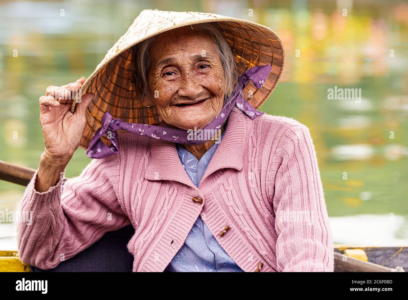 Old Vietnamese woman in violet sweater holds her hat while smiling Stock Photo