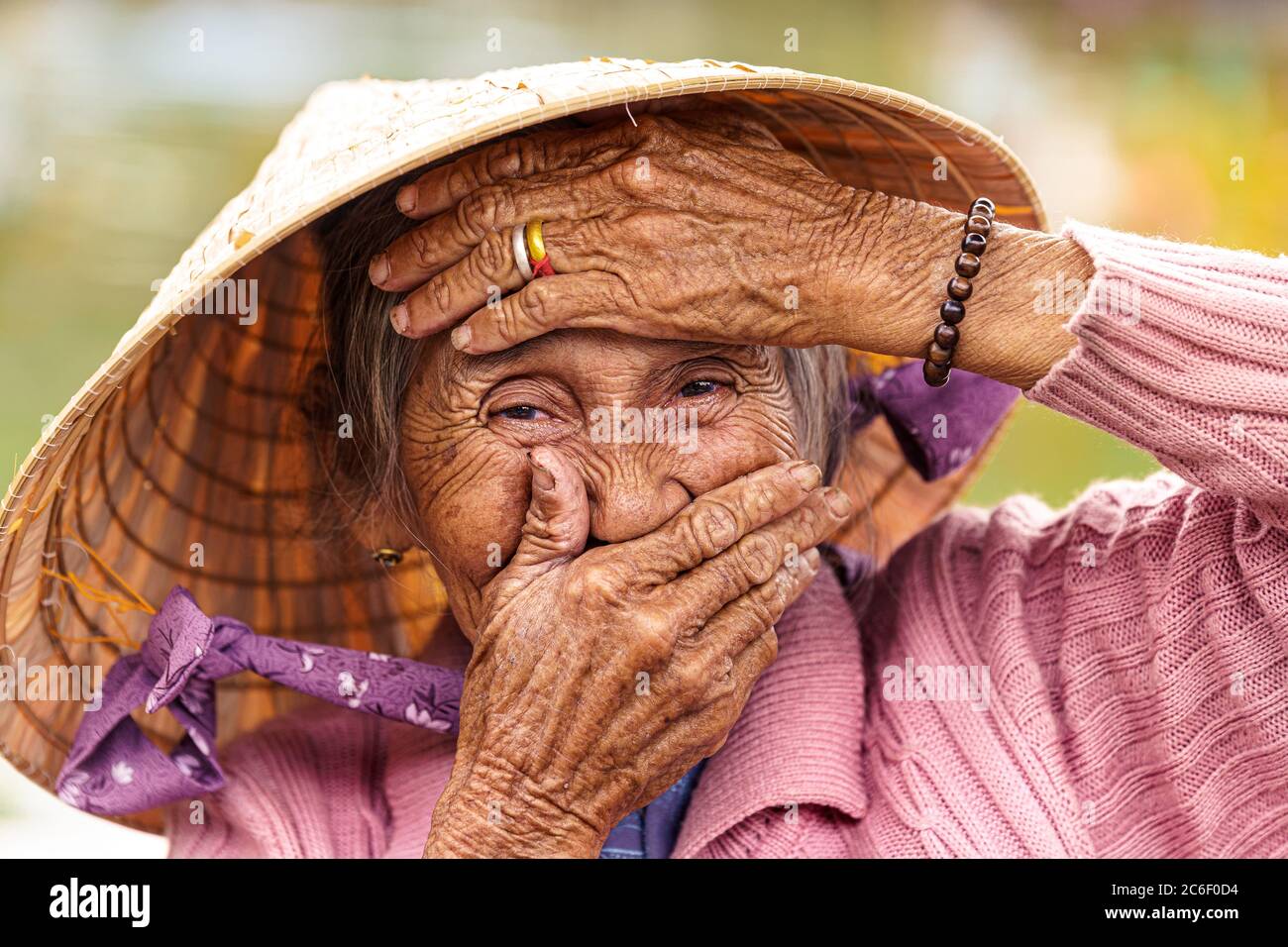 Old Vietnamese woman in violet sweater puts her hands to her face to cover her mouth and forehead. Stock Photo